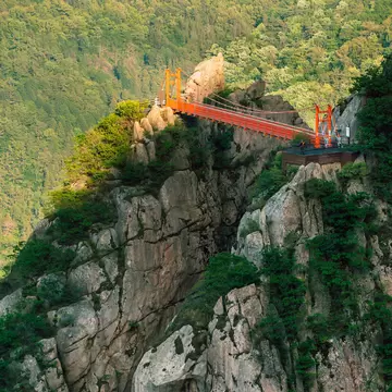 A red suspension footbridge between two rocky granite peaks above a forested valley in Wolchulsan National Park, South Korea.