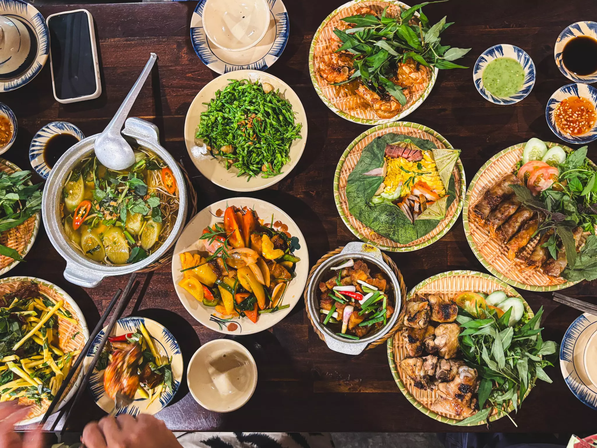 A variety of colorful dishes arranged on plates and in serving bowls. A hand with chopsticks is in the lower left corner.