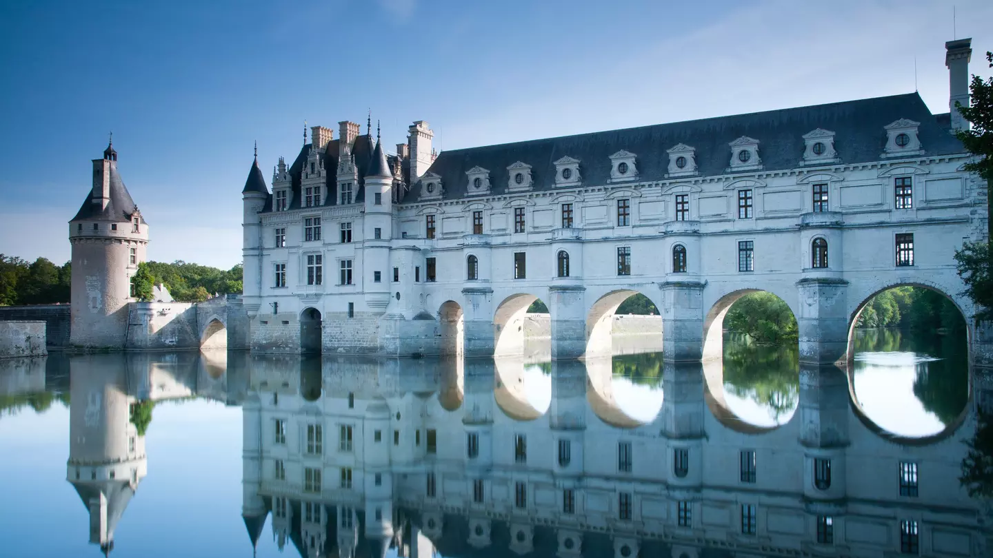 A large white castle-like building with large arches underneath it is built over a river