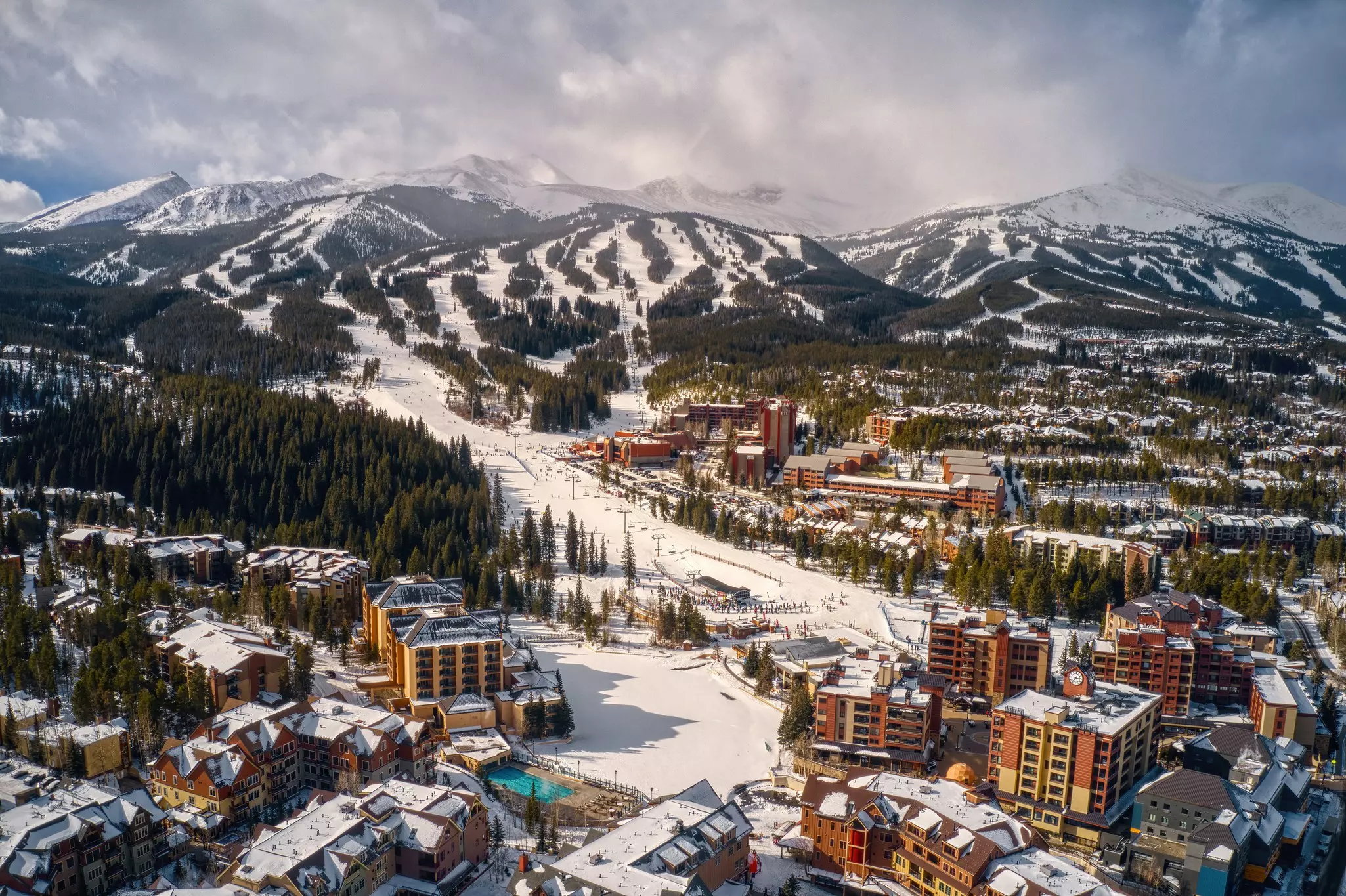 An aerial view of a snow-covered ski town at the base of several skiing mountains.