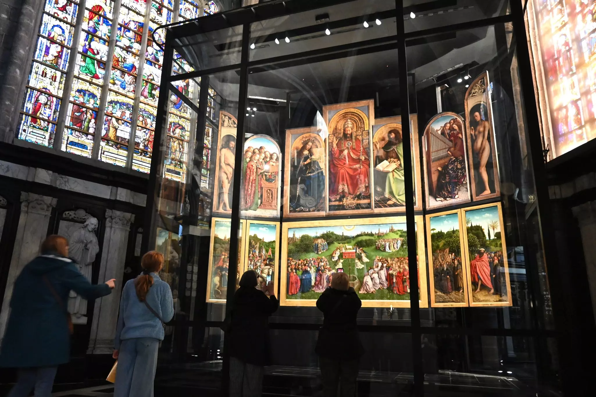 People look at a giant painted altarpiece, on display behind glass, in a side chapel of a church.