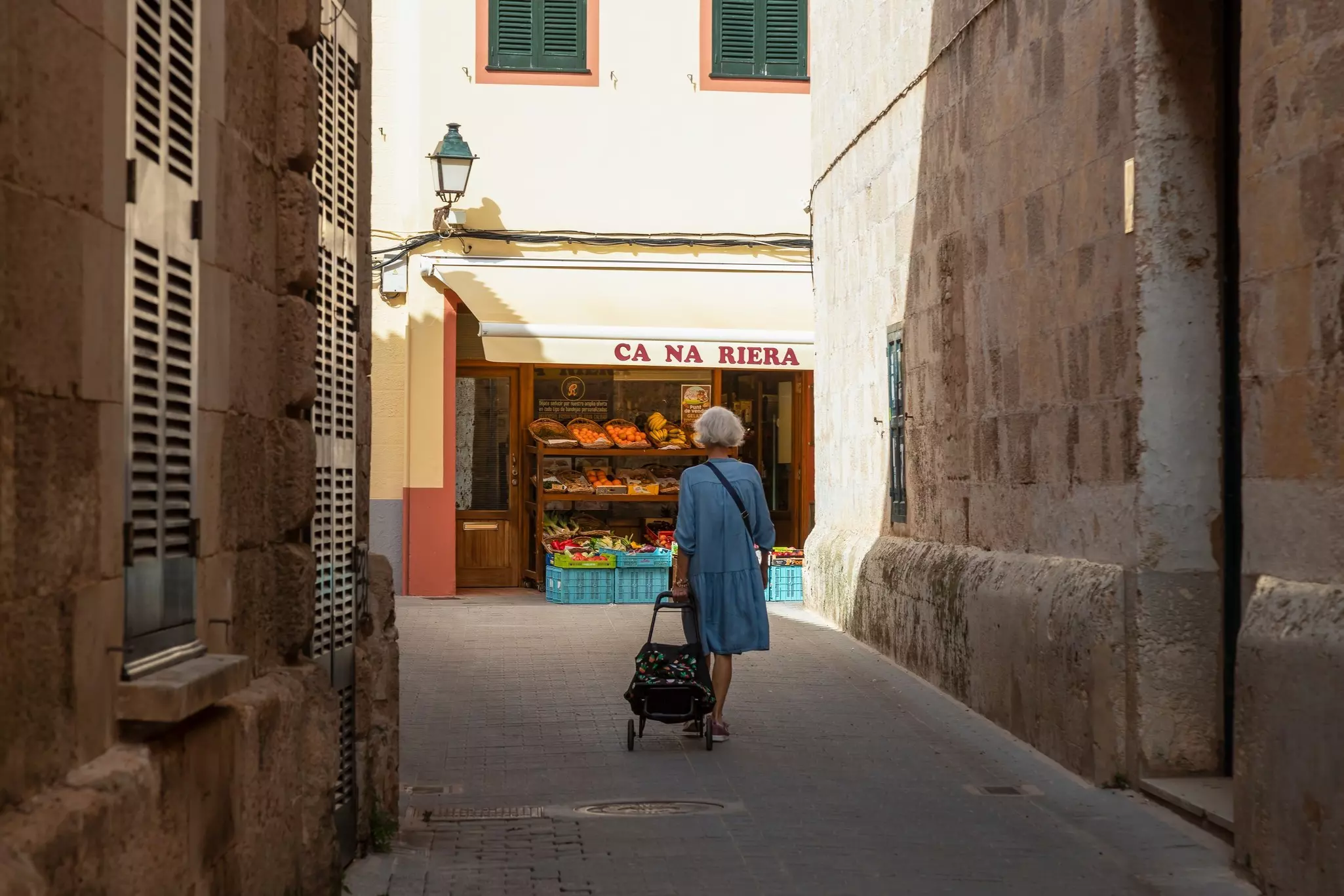 A woman walks with a shopping bag on wheels past a greengrocer.