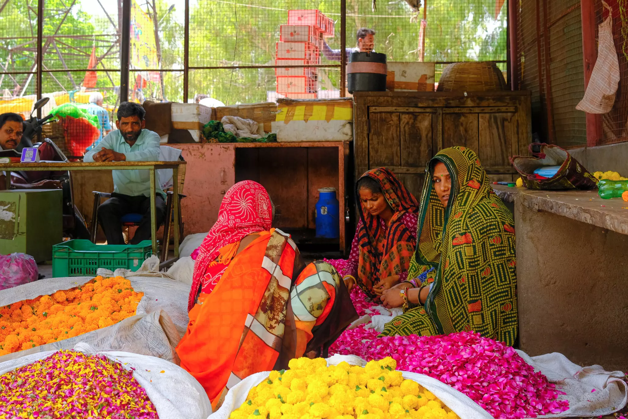 Three women dressed in saris with head covereds sit surrounded by bags of colorful flowers on petals at a market.