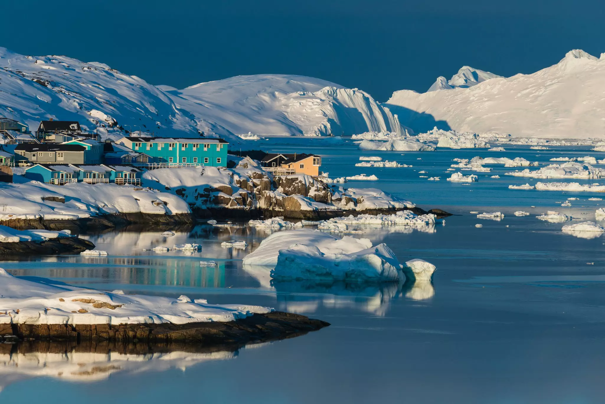 Low buildings painted in green, turquoise and yellow in a snowy landscape by a body of water with icebergs floating on the surface.