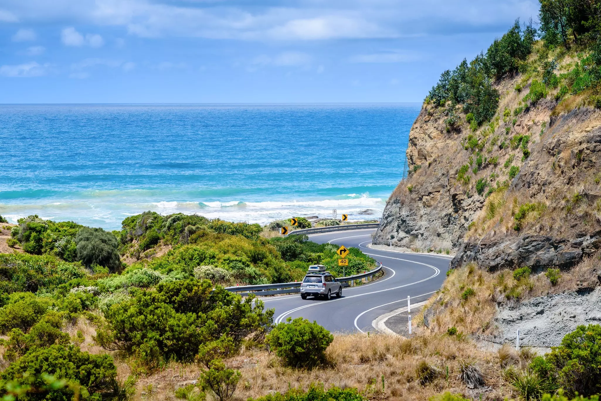 Cars drive on a windy road hugging the ocean next to cliffs
