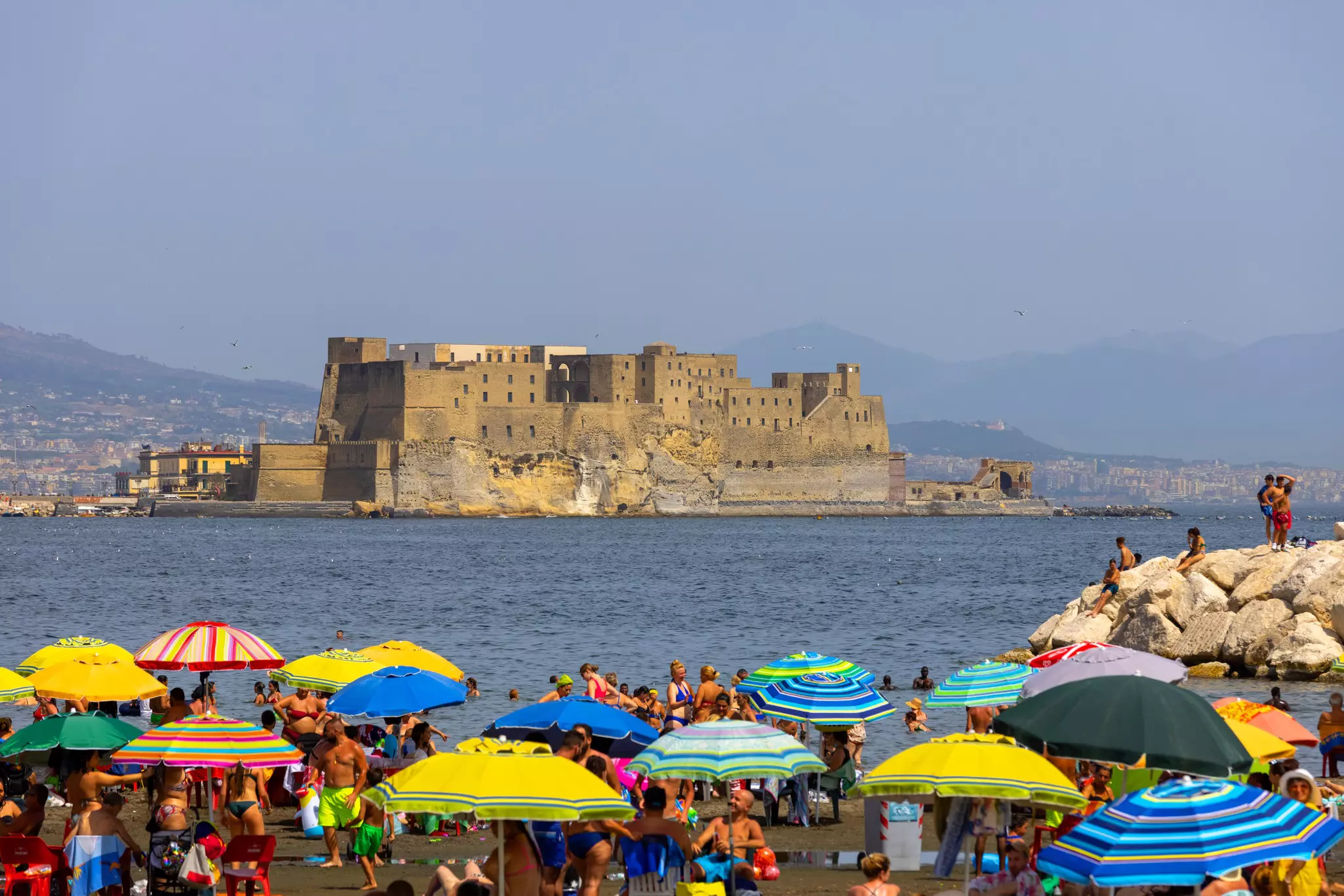 People relaxing on the Mappatella Beach by the Tyrrhenian Sea next to the boulevard Chiaia, colorful umbrellas, Castel dell'Ovo in the background