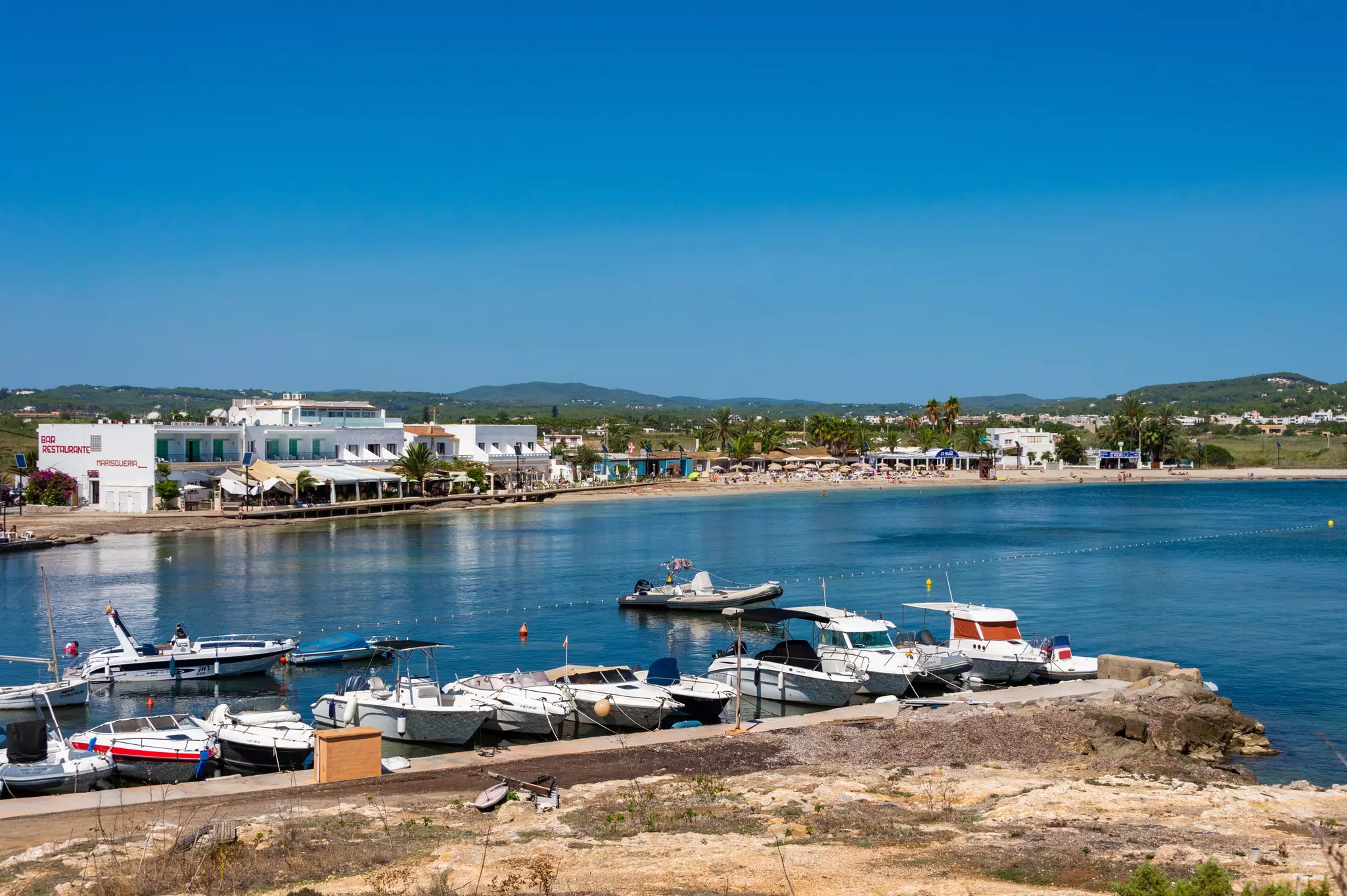 Small boats docked on the brilliant blue waters of a beach. Low-profile buildings line the shore in the background