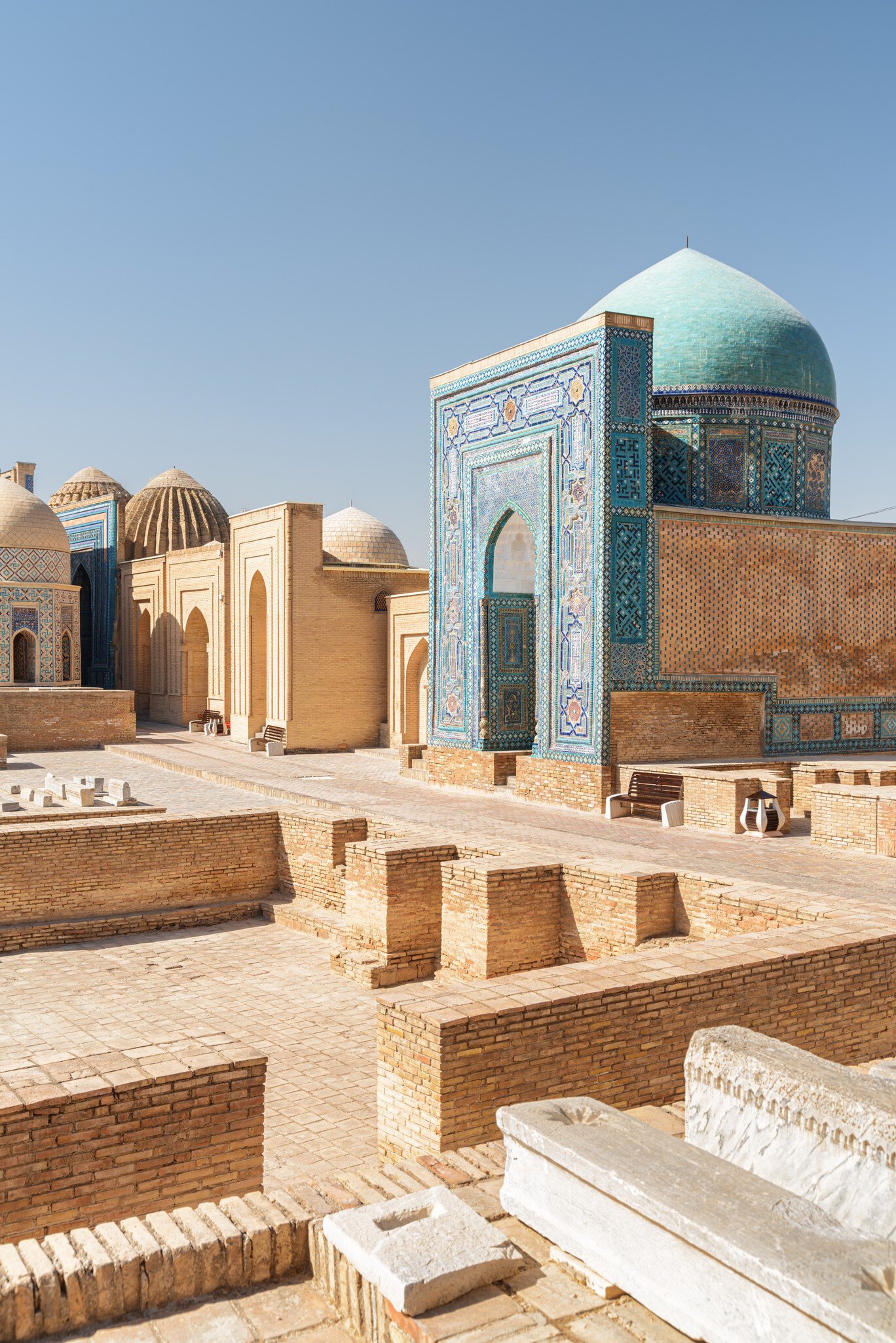 A mausoleum with a blue mosaic-tiled portal and turquoise dome beside tan brick tombs and domes under a clear sky.