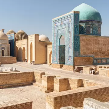 A mausoleum with a blue mosaic-tiled portal and turquoise dome beside tan brick tombs and domes under a clear sky.