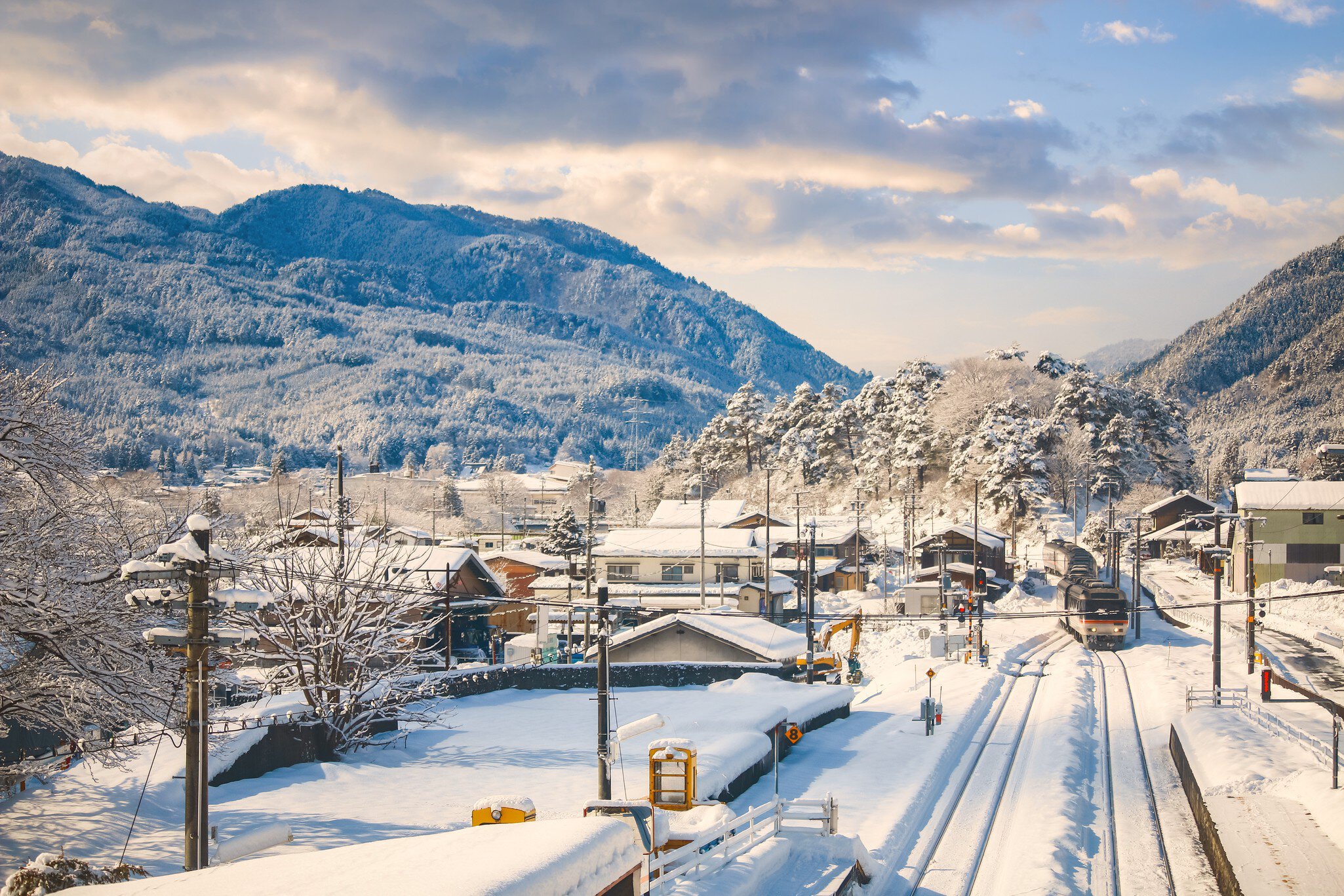 Train on snowy tracks beside snow-covered rooftops and forested mountains in Takayama, Japan, in winter