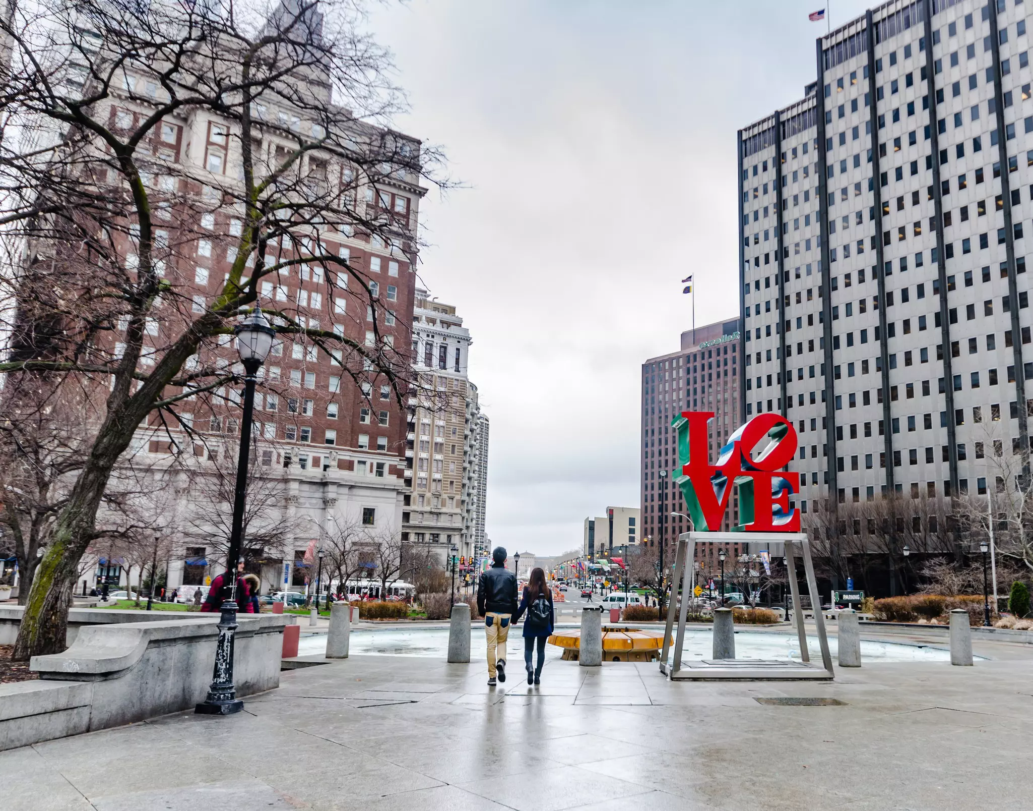 A couple walks hand in hand through a city plaza near a statue of letters reading "LO" above and "VE"below in red on an open metal base with city buildings in the distance on a cloudy day.
