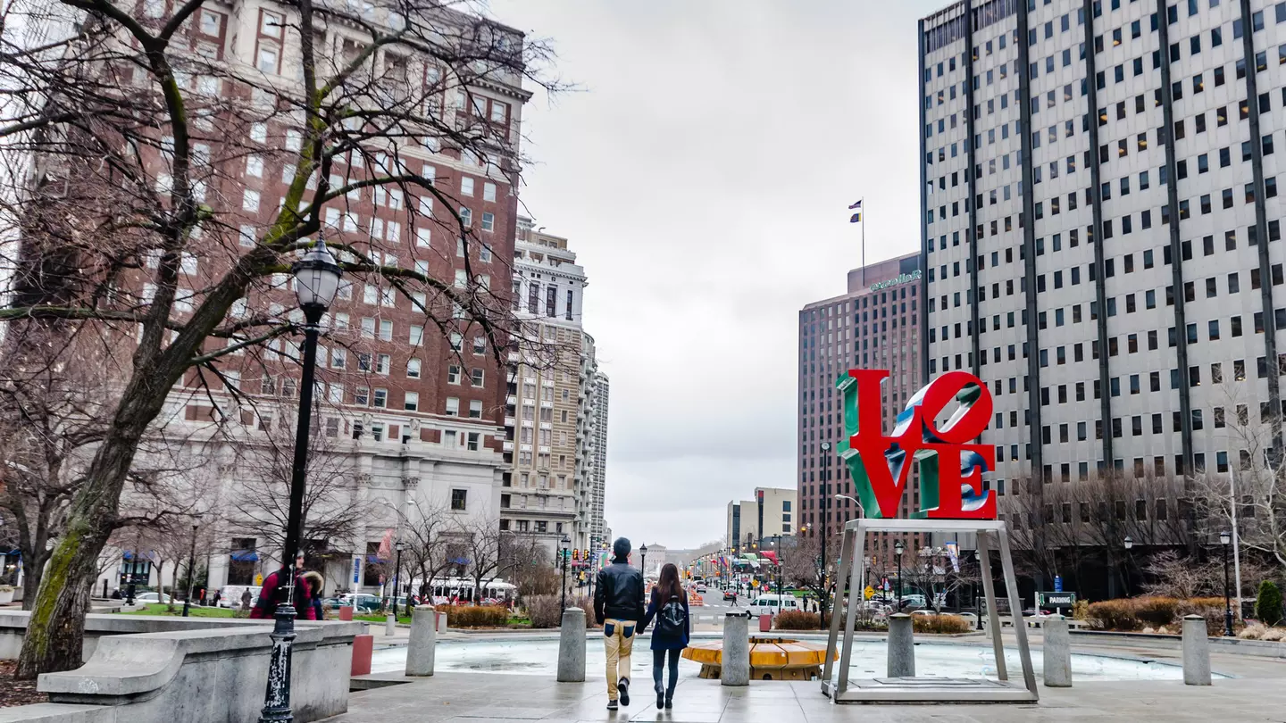 A couple walks hand in hand through a city plaza near a statue of letters reading "LO" above and "VE"below in red on an open metal base with city buildings in the distance on a cloudy day.