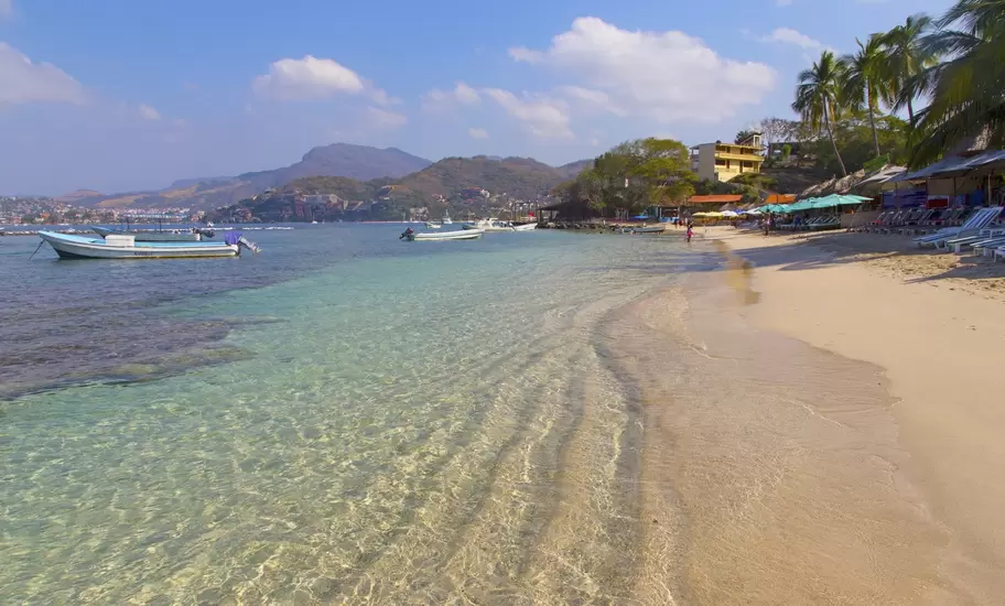 Clear turquoise water lapping a sandy beach with moored boats, palapa umbrellas and green hills in the distance.