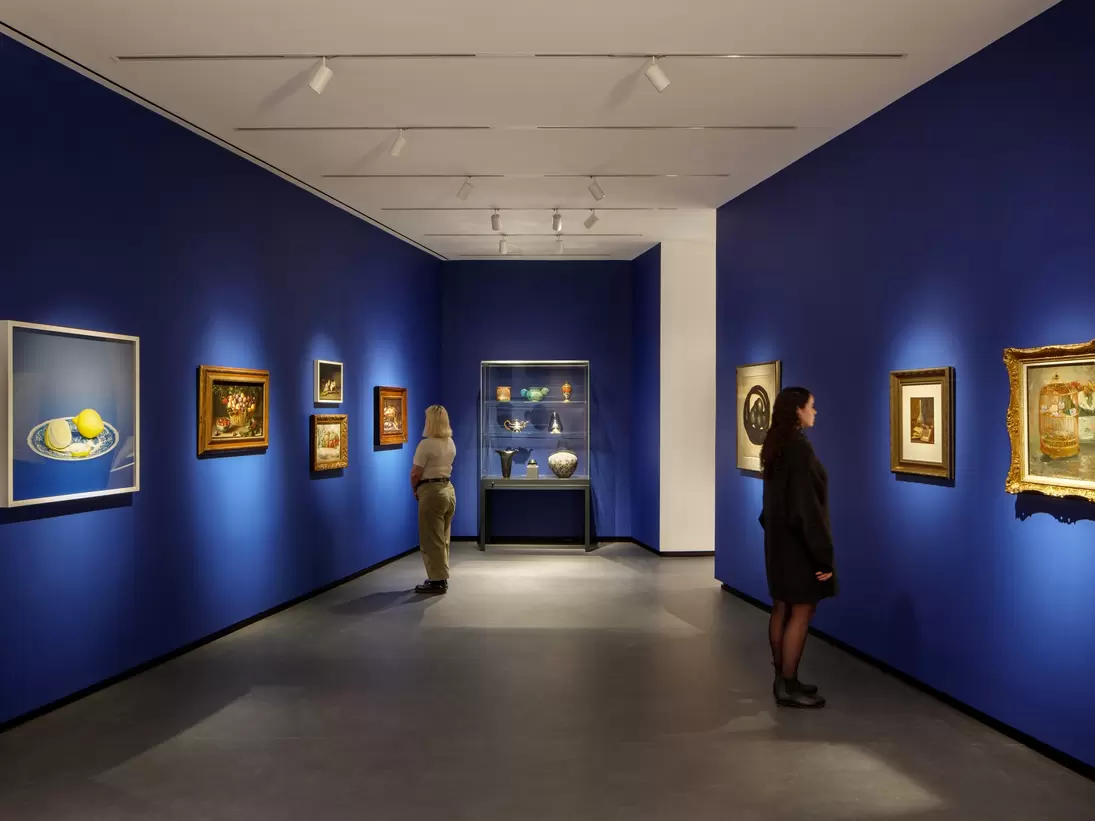 Museum visitors observe artworks hanging on the royal blue walls of a small gallery bay. Paintings of various sizes are on display, and small sculptures are in a glass case.