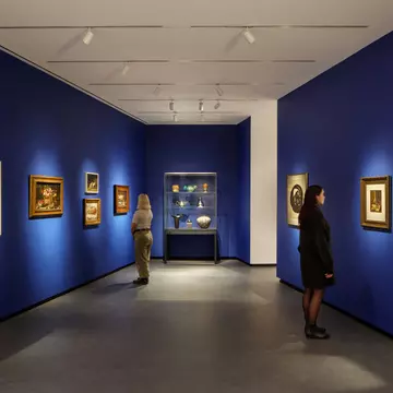 Museum visitors observe artworks hanging on the royal blue walls of a small gallery bay. Paintings of various sizes are on display, and small sculptures are in a glass case.
