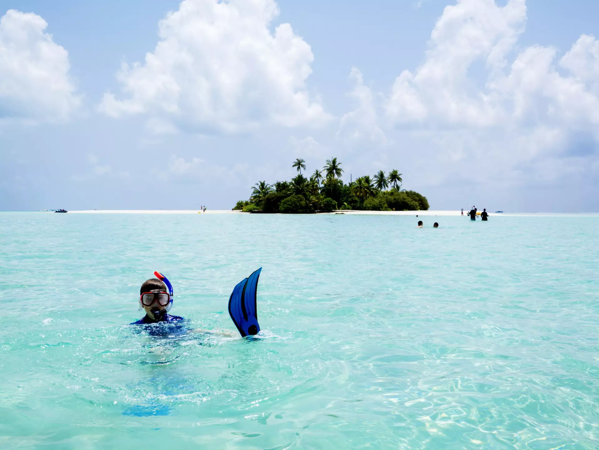 A child wearing a snorkeling mask and blue fins floats in turquoise water off a small island with white sand and greenery.