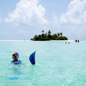 A child wearing a snorkeling mask and blue fins floats in turquoise water off a small island with white sand and greenery.