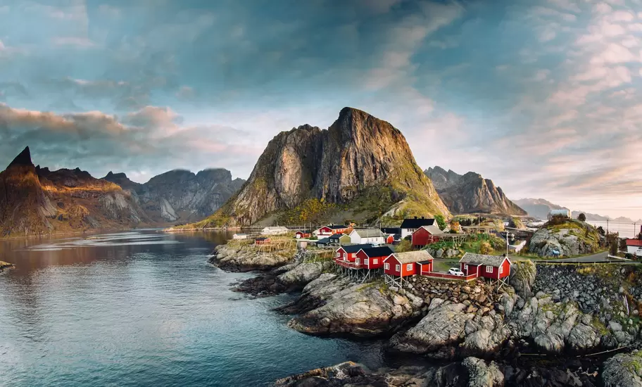 Red rorbu fishing cabins on rocky shore at Hamnøy in Lofoten, Nordland, beneath a sharp granite peak at dawn