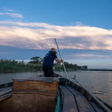 Man in a straw hat pushing a long pole from the bow of a wooden boat on a calm lagoon edged with reeds at dusk.