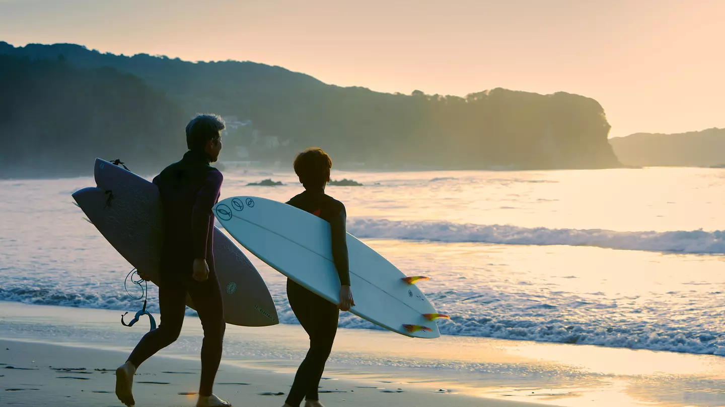 A man and woman head into the sea with their surfboards under their arms as the sun sets.