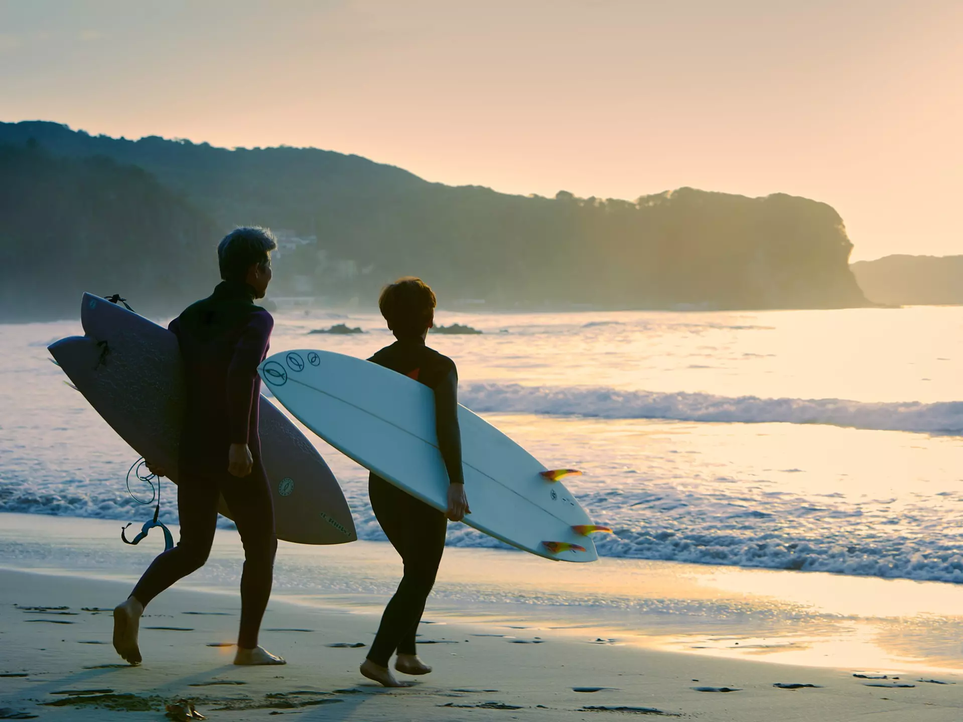 A man and woman head into the sea with their surfboards under their arms as the sun sets.