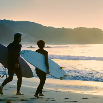 A man and woman head into the sea with their surfboards under their arms as the sun sets.