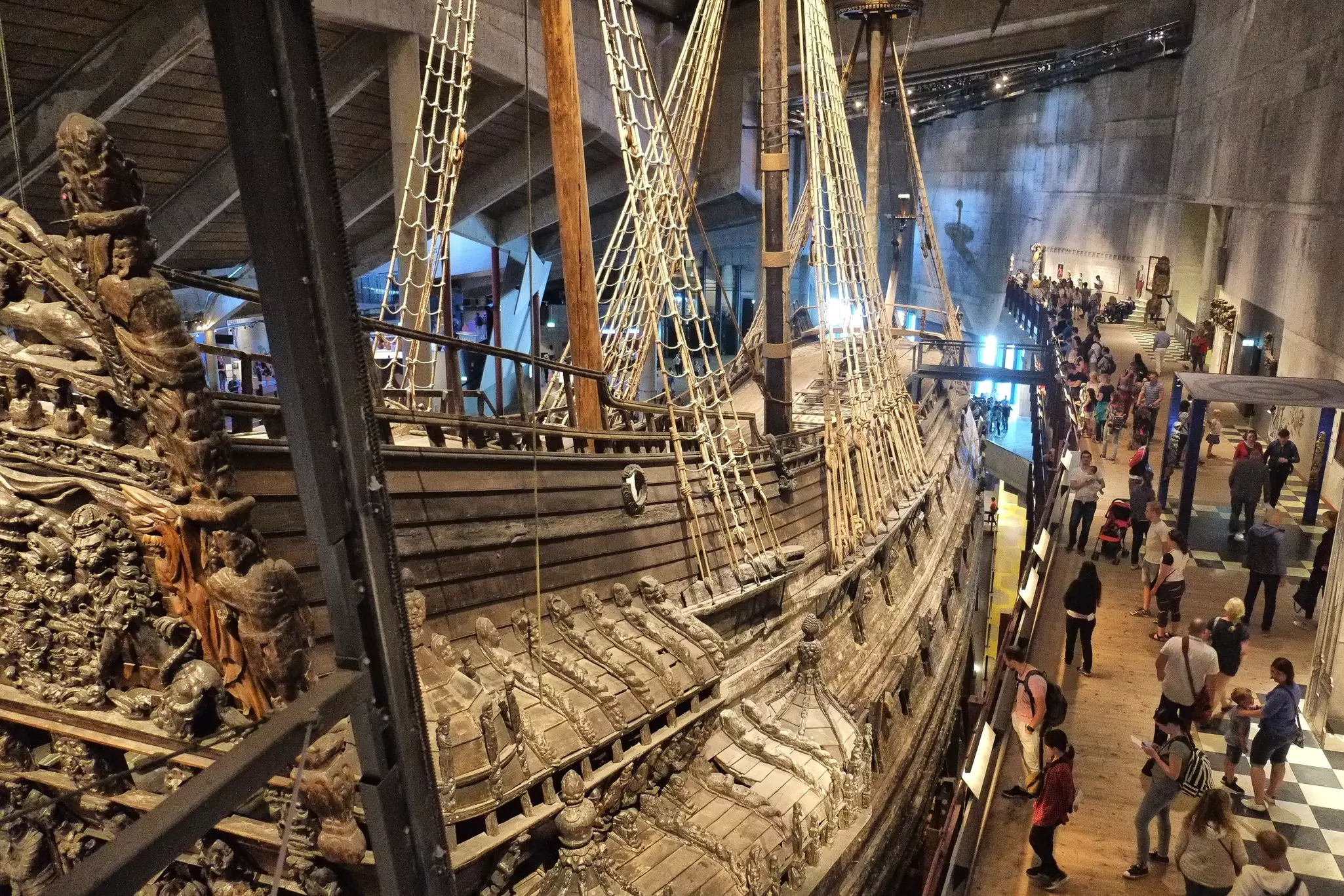 A huge wooden ship with ornate carvings over its sides stands within a museum as people walk on the nearby mezzanine. 