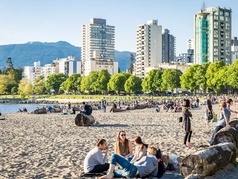 many people gathered on a beach in the sun a greenway in the background backs up to city buildings and mountains in the distance.