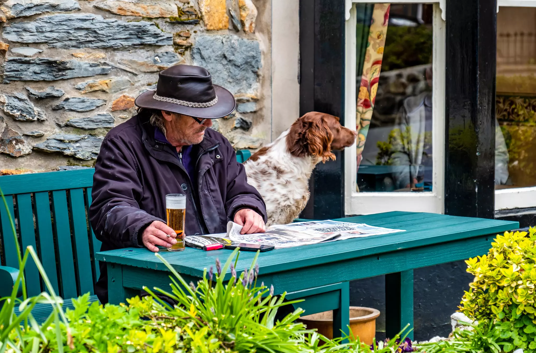 A man sits at an outdoor table at a pub, sipping a beer and reading a newspaper, with his dog.