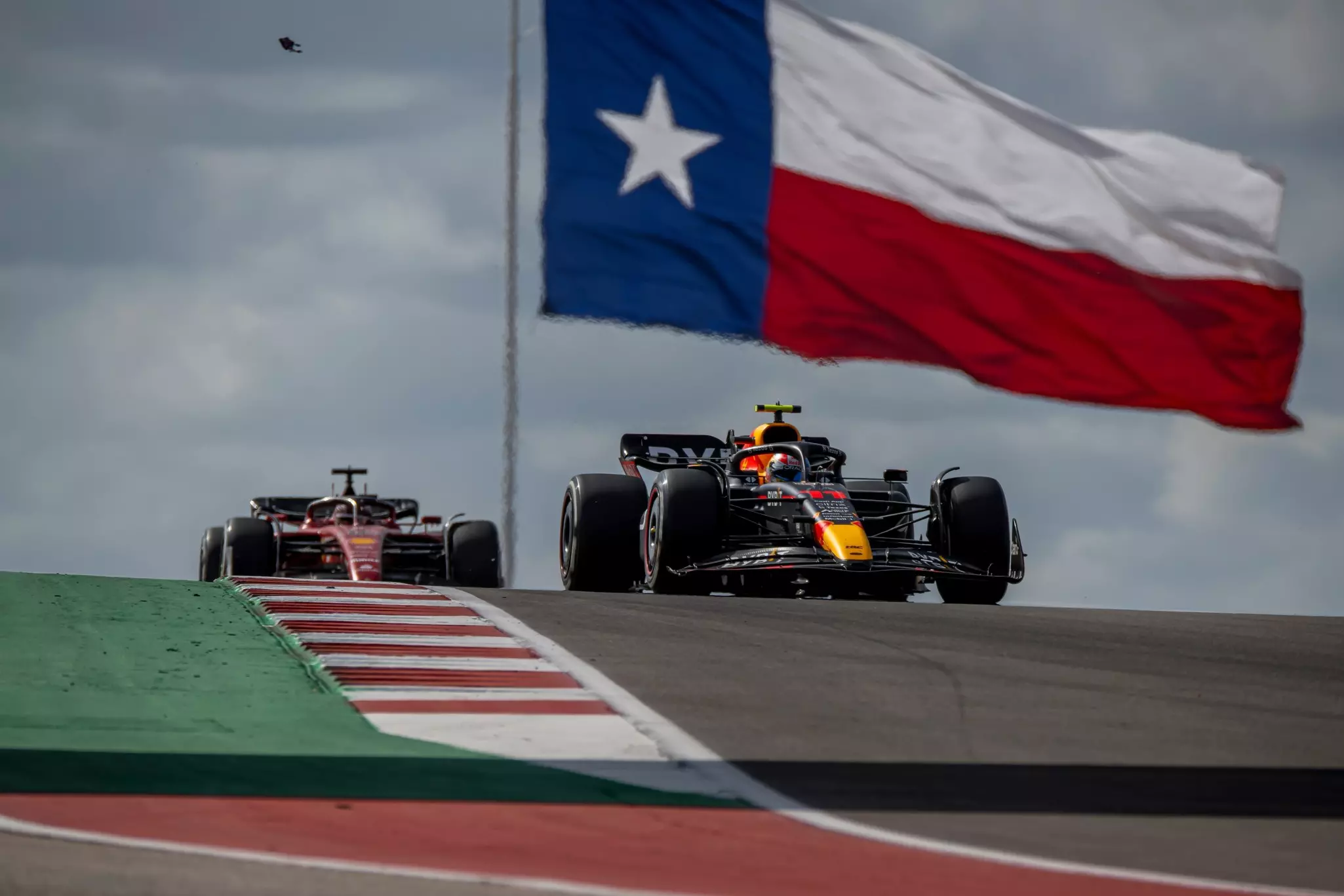 Two race cars driving on a track. A large red, white and blue flag with a star flies above