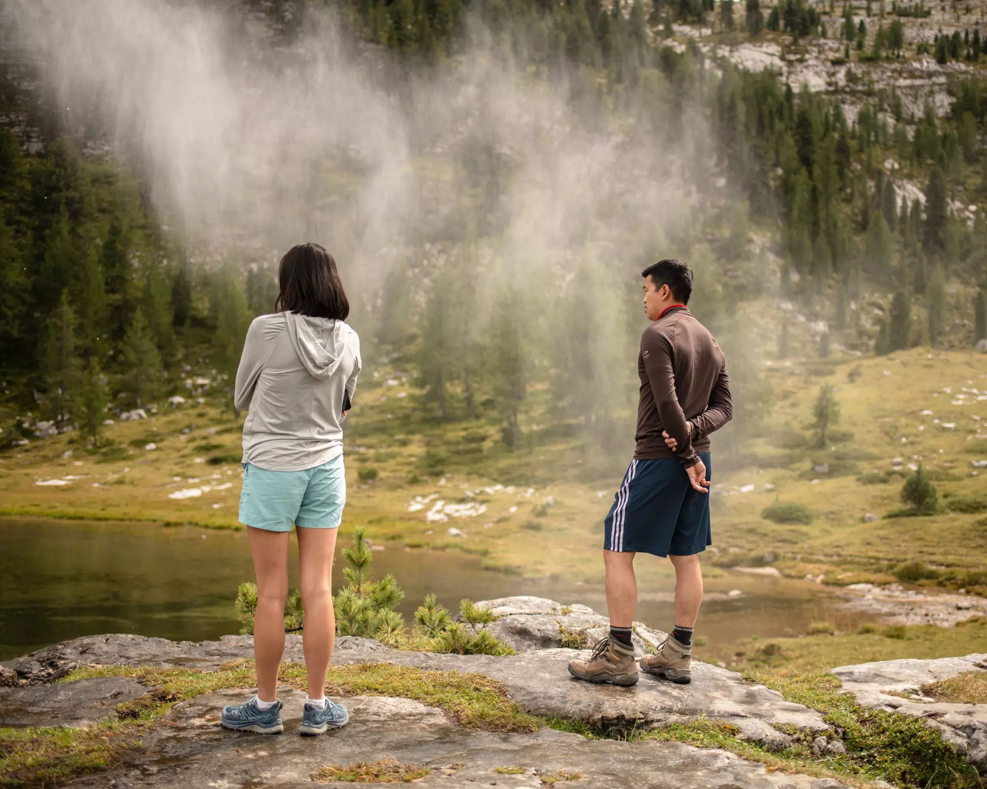 Two people in hiking clothes stand on a rock looking toward a lake in misty mountain wilderness in Italy.