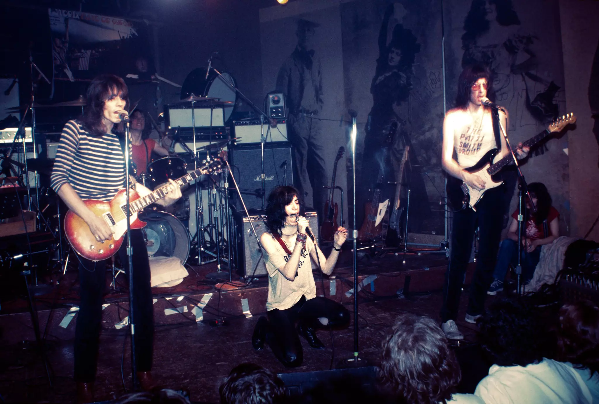 A woman kneels on a stage with a microphone wearing a white tshirt two people flank her playing the guitar a drums and speakers behind her.