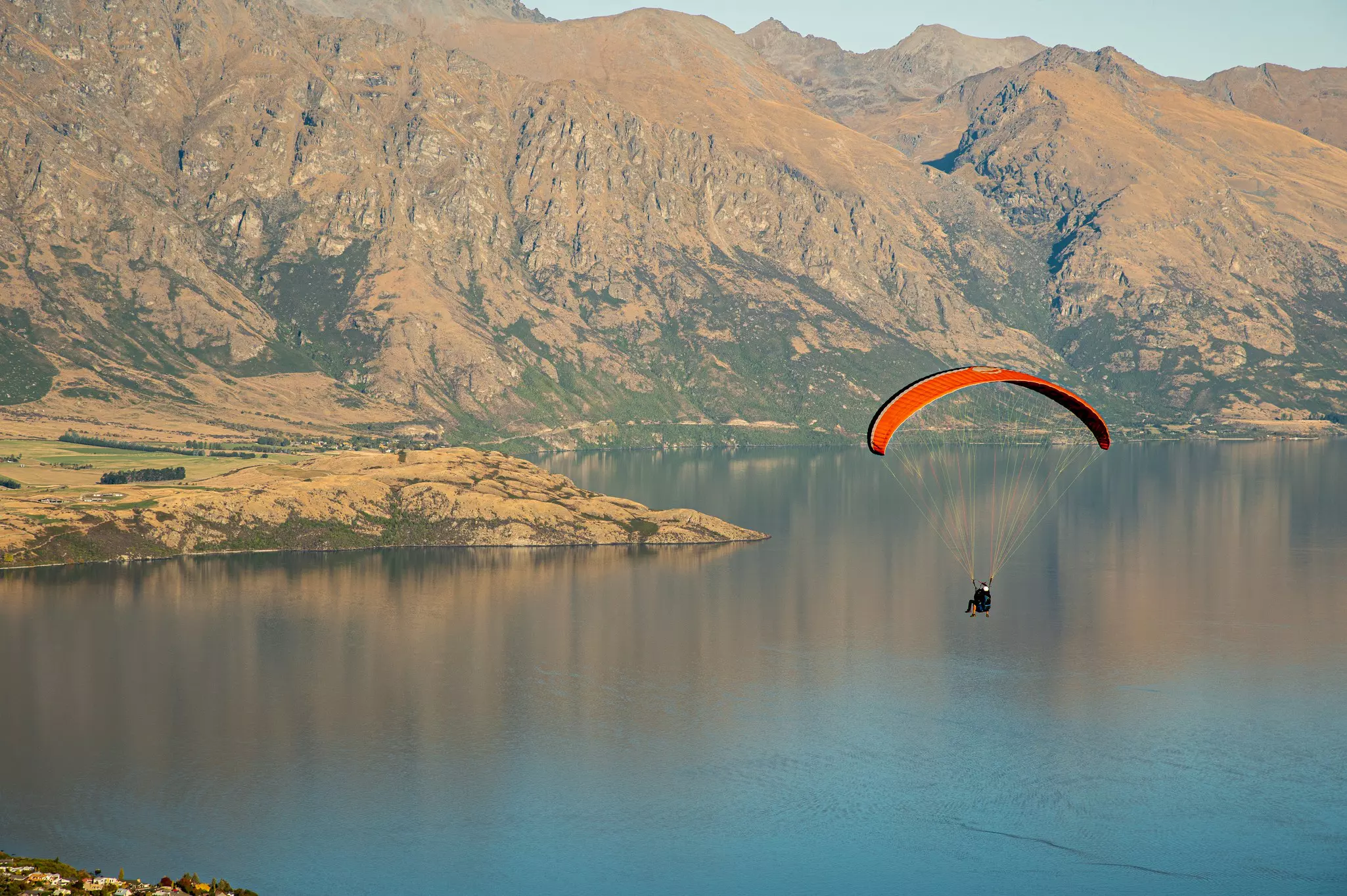 A paraglider with a red sail soars over a lake, with dramatic mountain scenery in the distance.