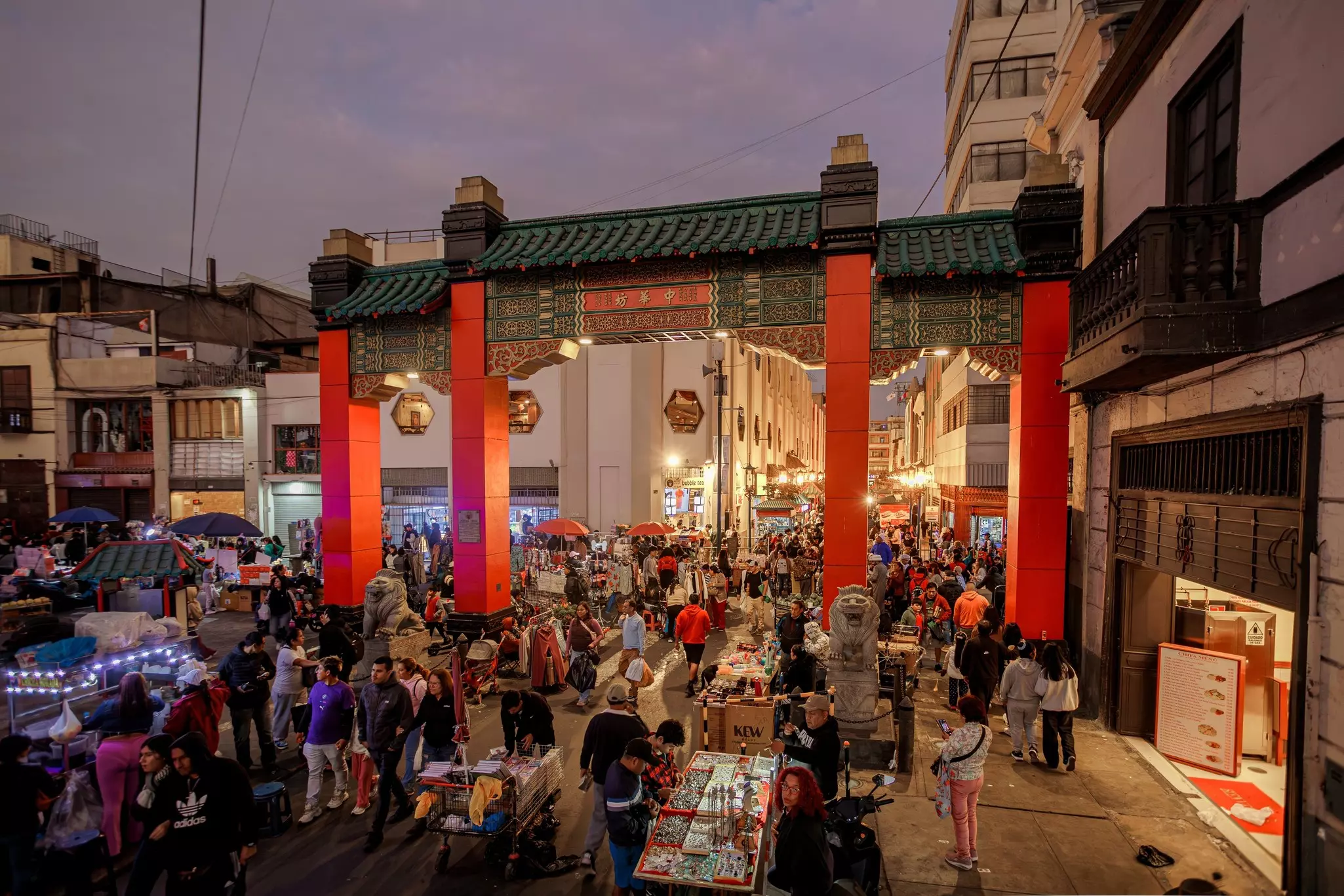 People in Lima, Peru, walk through a Chinese-style arch with red pillars and a green keystone at night.