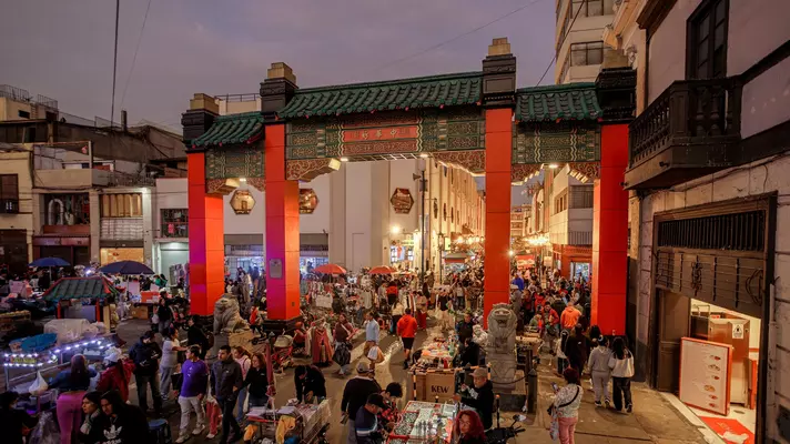People in Lima, Peru, walk through a Chinese-style arch with red pillars and a green keystone at night.