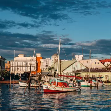 Painted wooden sailboats at moorings on dark rippled water in evening sun, backed by low pastel buildings under a blue sky.