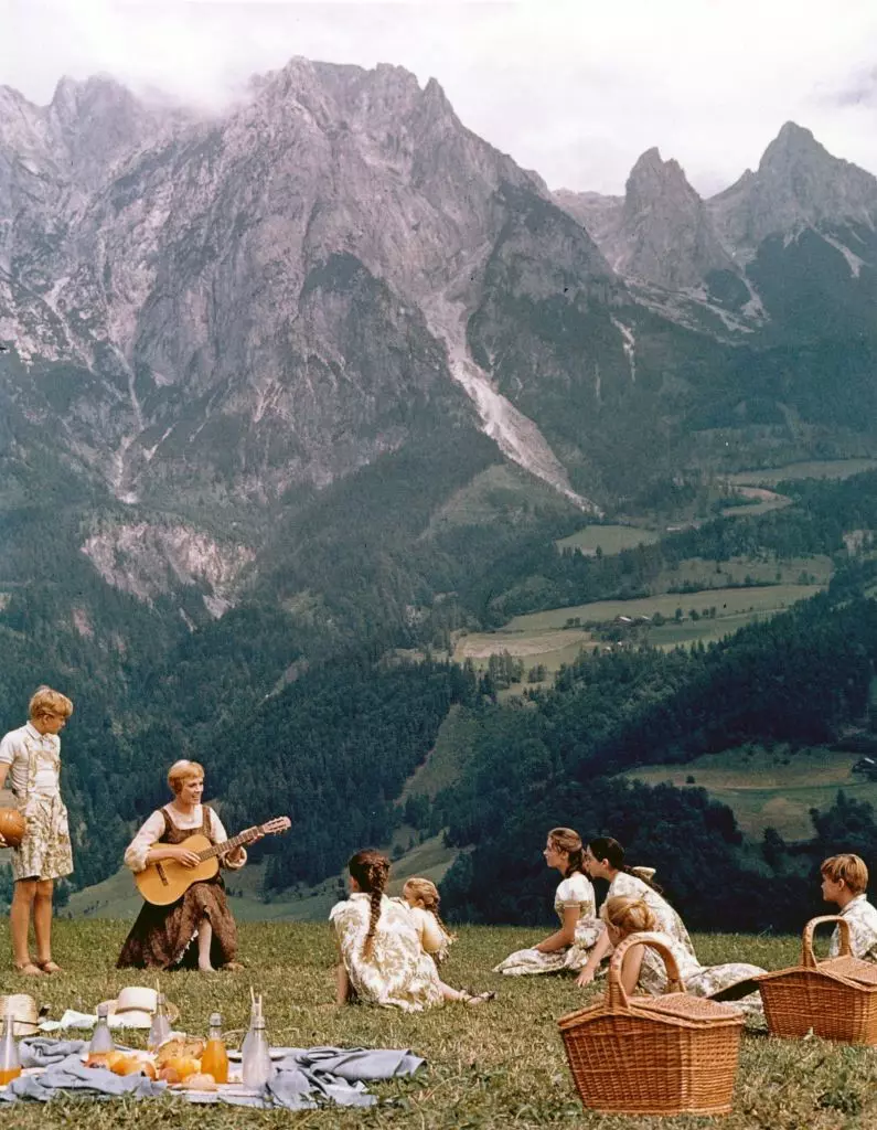 A woman with a guitar plays and sings to a small group of children sat on the grassy meadows in a mountainous region