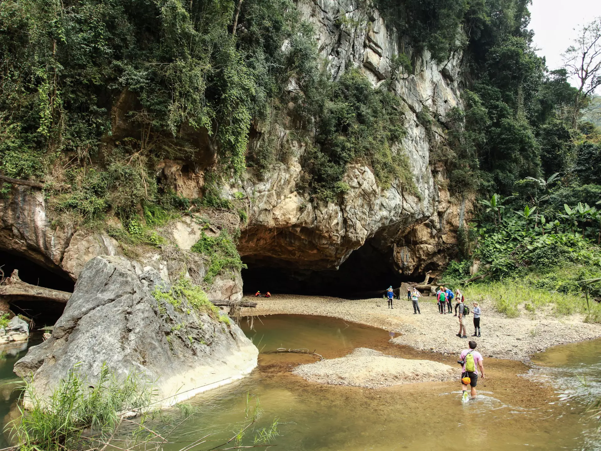 People wade through a river on their approach to a cave mouth surrounded by jungle.