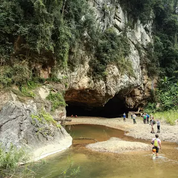 People wade through a river on their approach to a cave mouth surrounded by jungle.