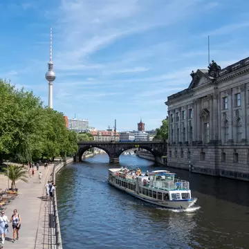 A tourist boat travels down a river below a tall TV tower: Museumsinsel (Museum Island), in Mitte, Berlin.
