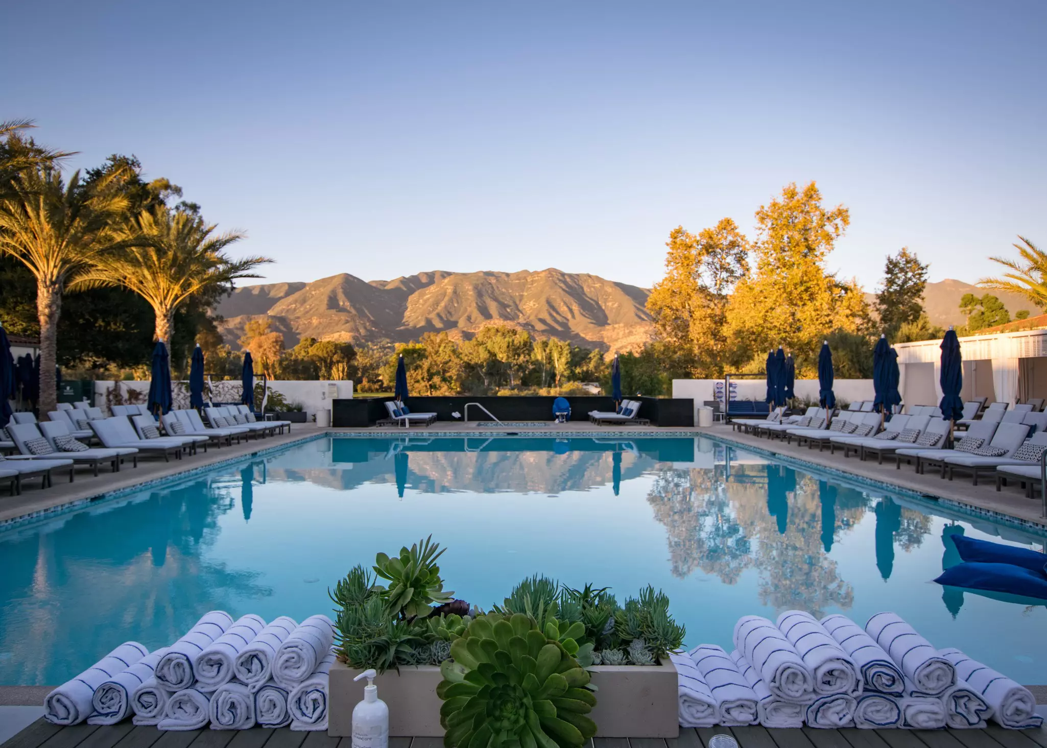 View of a swimming pool surrounded by lounge chairs with rolled towels in the foregroun and mountains in the distance.