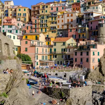 Colourful tower houses climbing a rocky hillside in Manarola above a small boat ramp, with people sunbathing on rocks.