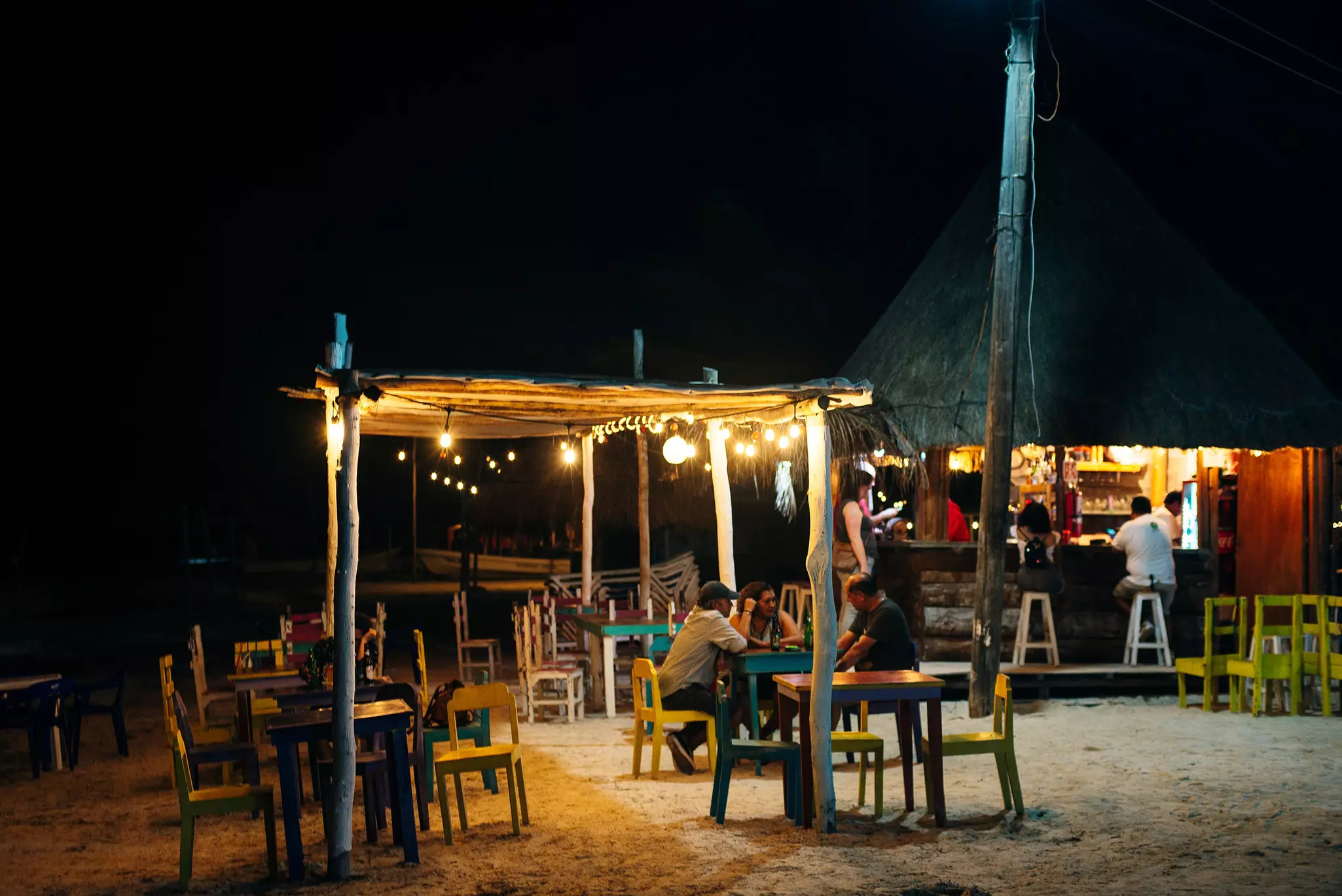 People sit at outdoor tables by night a bar on the beach in a tropical destination